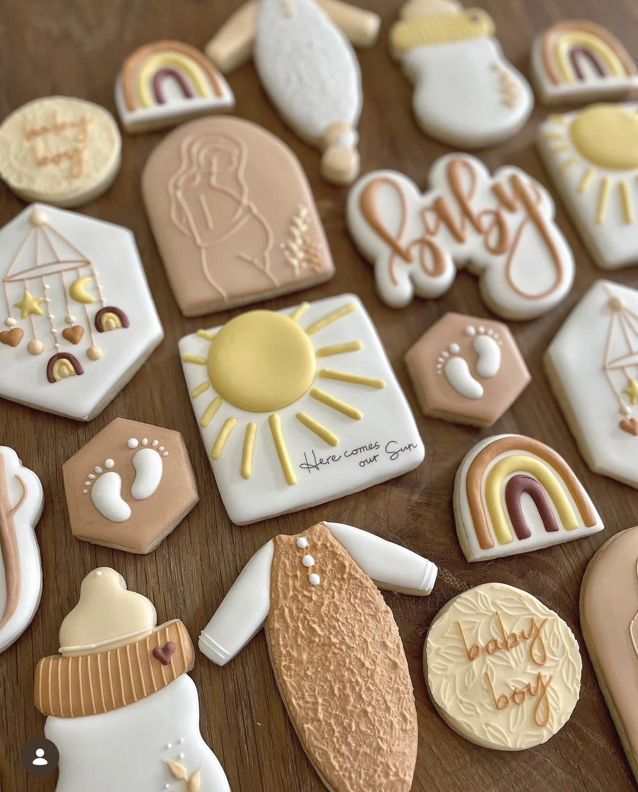 Assorted decorated baby shower sugar cookies on a wooden table, featuring rainbows, sunshine, baby footprints, a pregnant woman, baby clothes, and "Here comes our Sun" lettering.