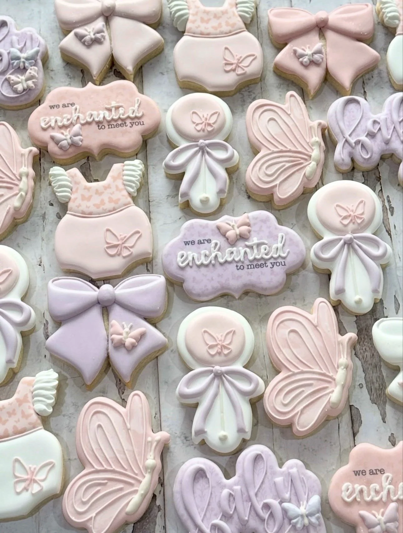 Pastel decorated sugar cookies featuring butterflies, bows, baby outfits, angels, and the phrase “we are enchanted to meet you”
