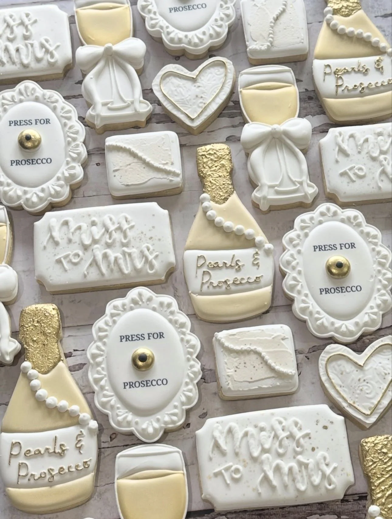 Assorted bridal shower cookies decorated as champagne bottles, glasses, hearts, wedding dresses, and prosecco press buttons in white, cream, and gold.