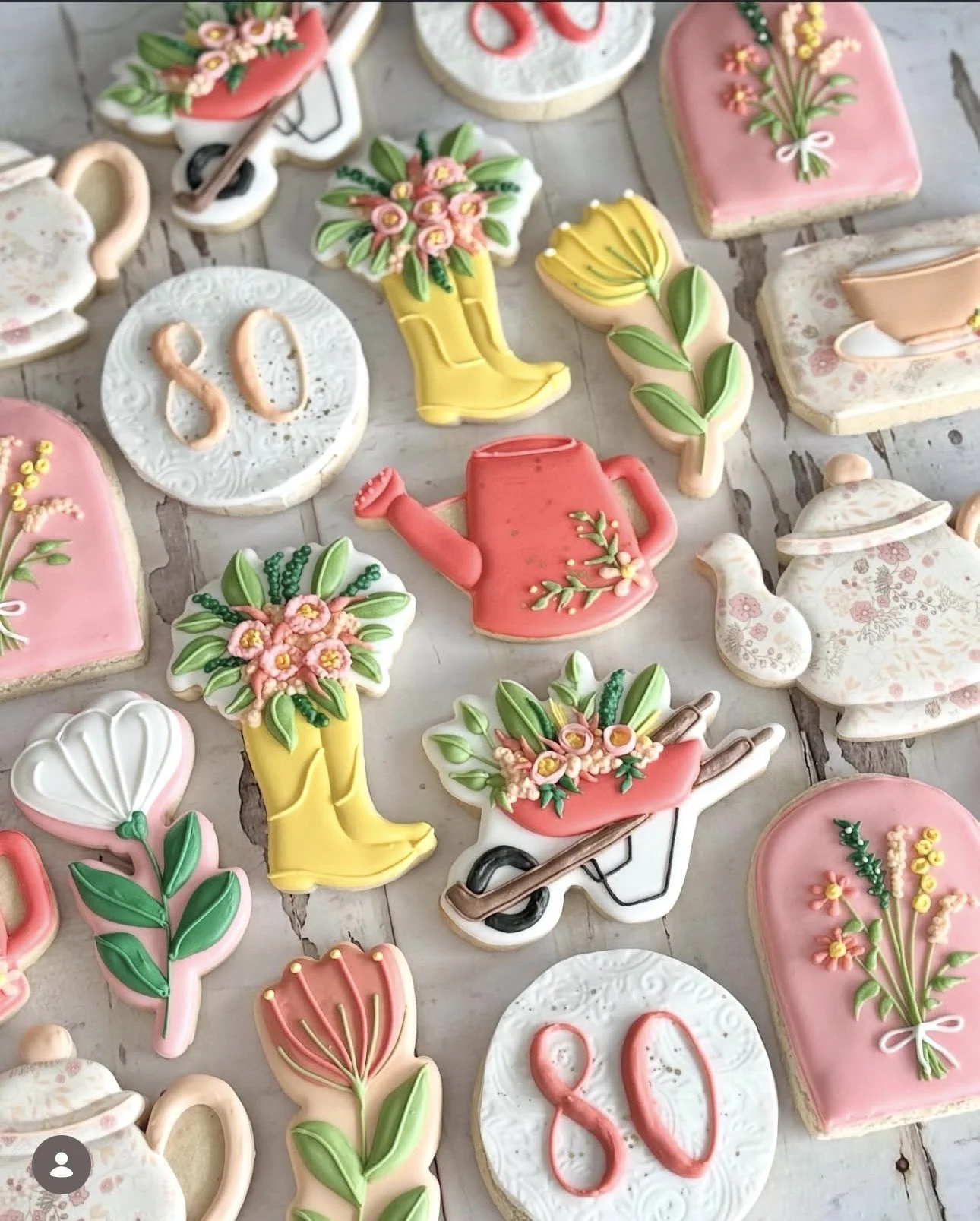 Colorful floral cookies shaped like gardening tools, watering cans, boots, flowers, and teacups arranged on a rustic wooden table