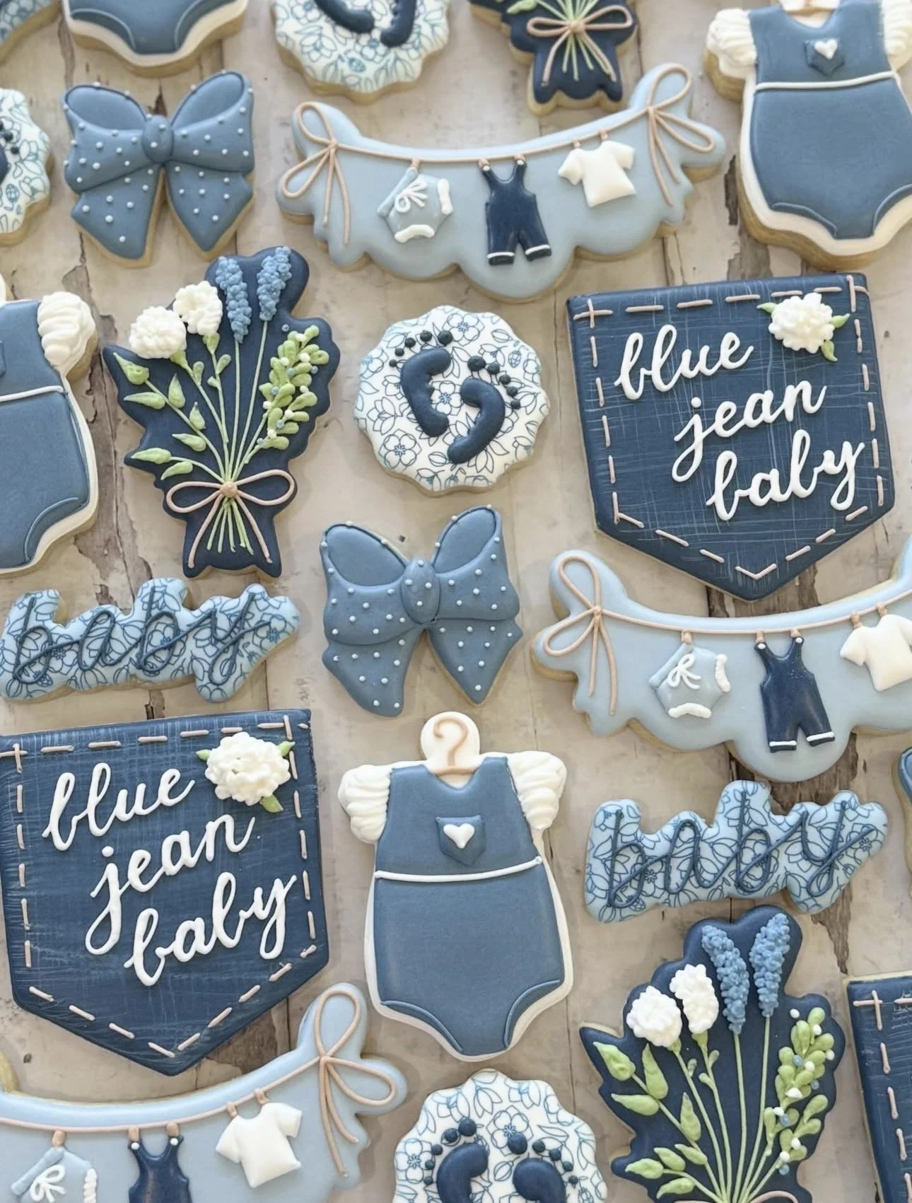 Blue-themed baby shower cookies with baby clothes, bows, floral bouquets, onesies, and signs reading “blue jean baby.”