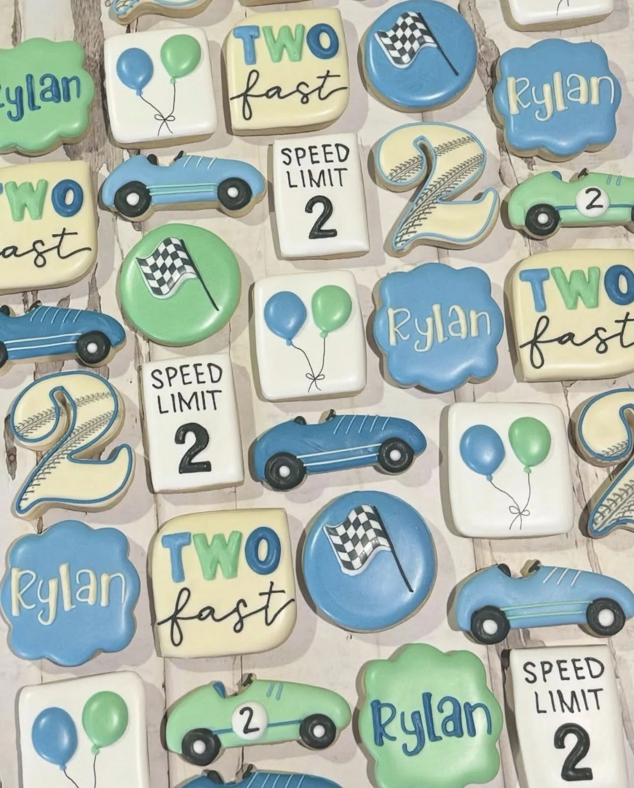 Decorated race car-themed cookies with checkered flags, vintage cars, balloons, baseball-style number twos, and personalized “Rylan” and “Two fast” lettering.