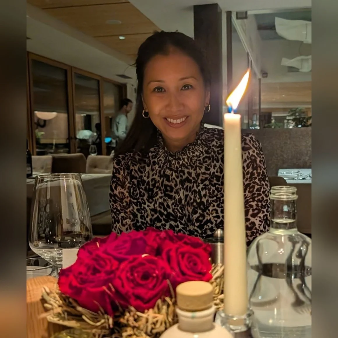 A woman with dark hair and earrings smiling at a dinner table with a candle, a bouquet of red roses, a glass of white wine, and a water bottle in a restaurant setting.
