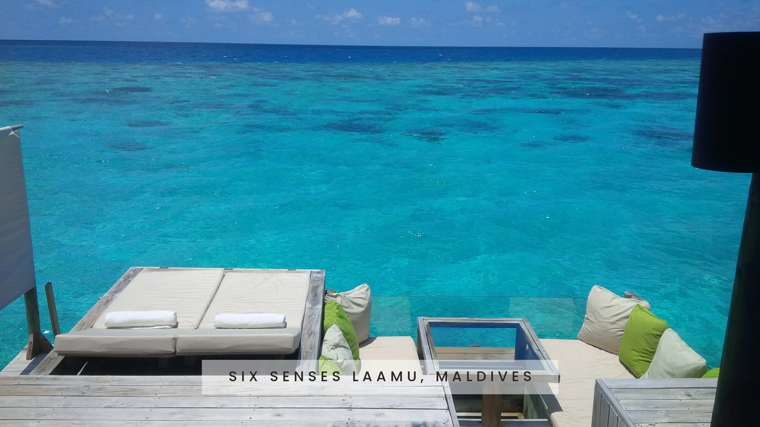 View of outdoor lounge chairs and cushions near the ocean with blue water and sky in Lamu, Maldives.