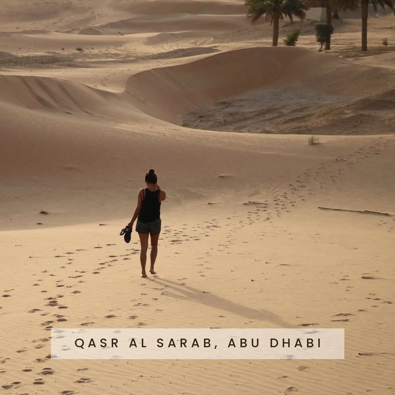 A woman walking barefoot in a desert with sand dunes in Qasr al Sarab, Abu Dhabi, carrying her shoes, with palm trees in the background.