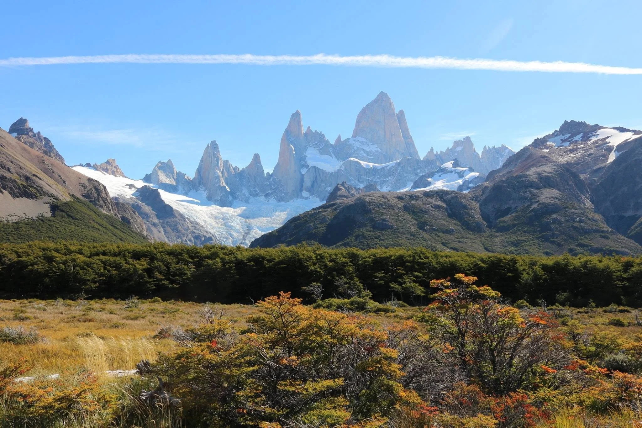 Snow-capped mountain peaks with lush green foliage and colorful autumn trees in the foreground under a clear blue sky.