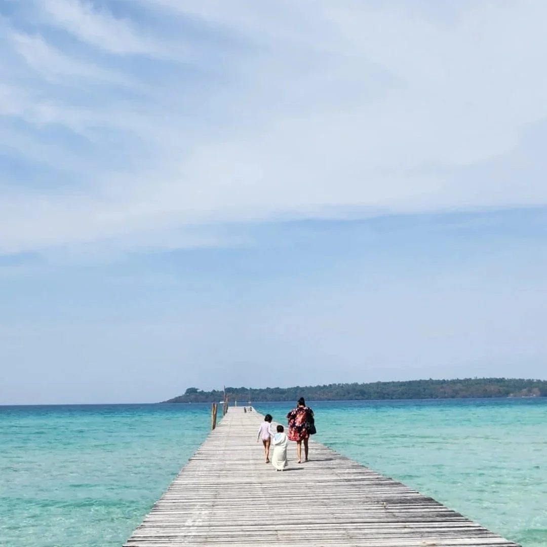 People walking on a wooden pier over turquoise water toward a distant island under a partly cloudy sky.