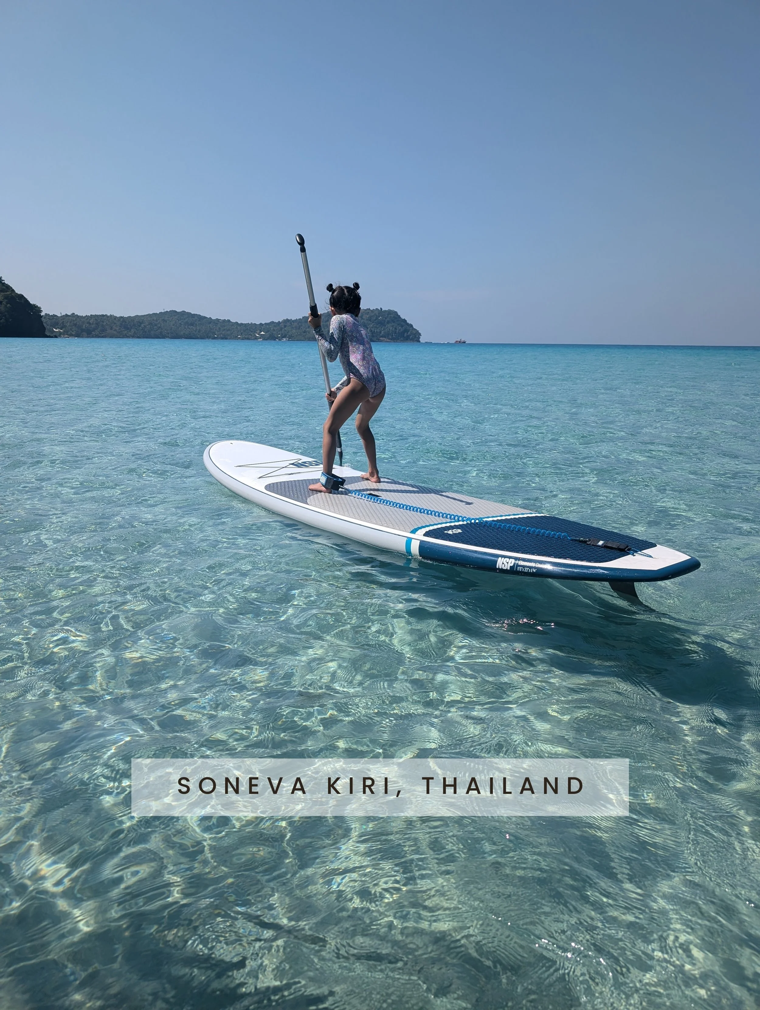 A girl with two buns hairstyle wearing a rainbow-colored swimsuit balancing on a paddleboard in clear shallow water near Soneva Kiri, Thailand, with a distant landmass and blue sky in the background.