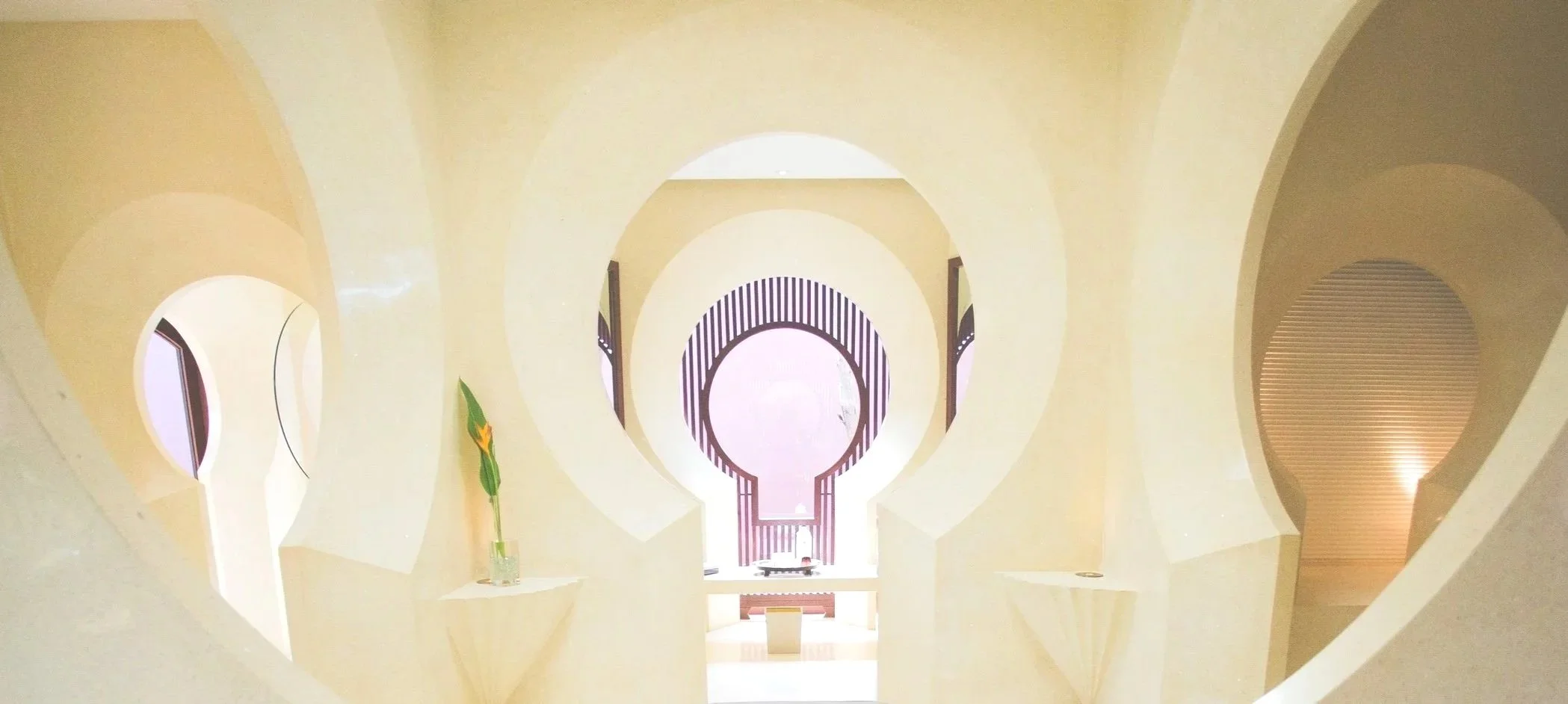 Interior view of a building with circular arches and modern architectural design, featuring a staircase, decorative wall, and a single flower in a vase.