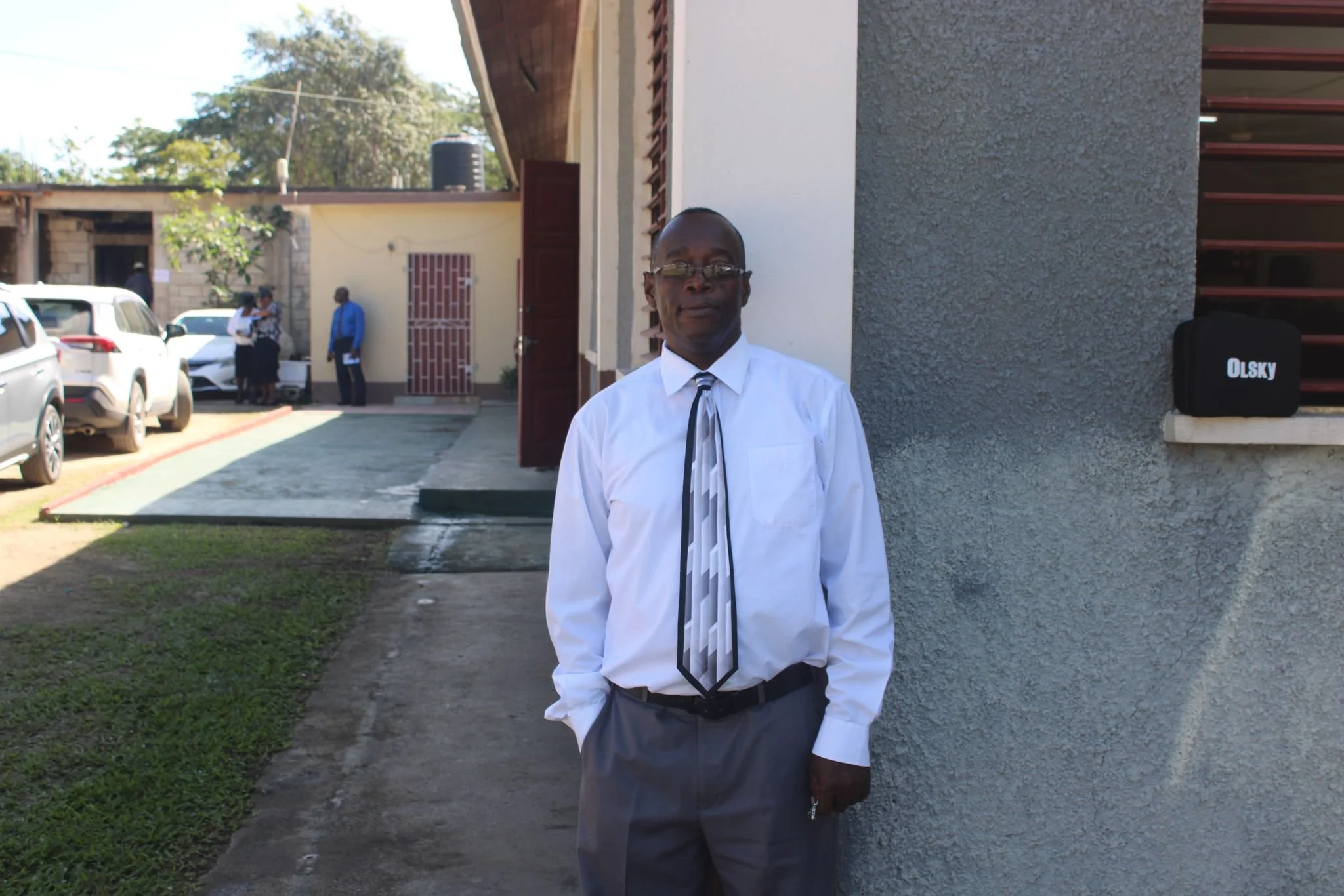 A man in a white shirt, striped tie, and gray pants stands outside near a building with gray and white walls. Background shows a parking lot with several cars, a few people, and trees.
