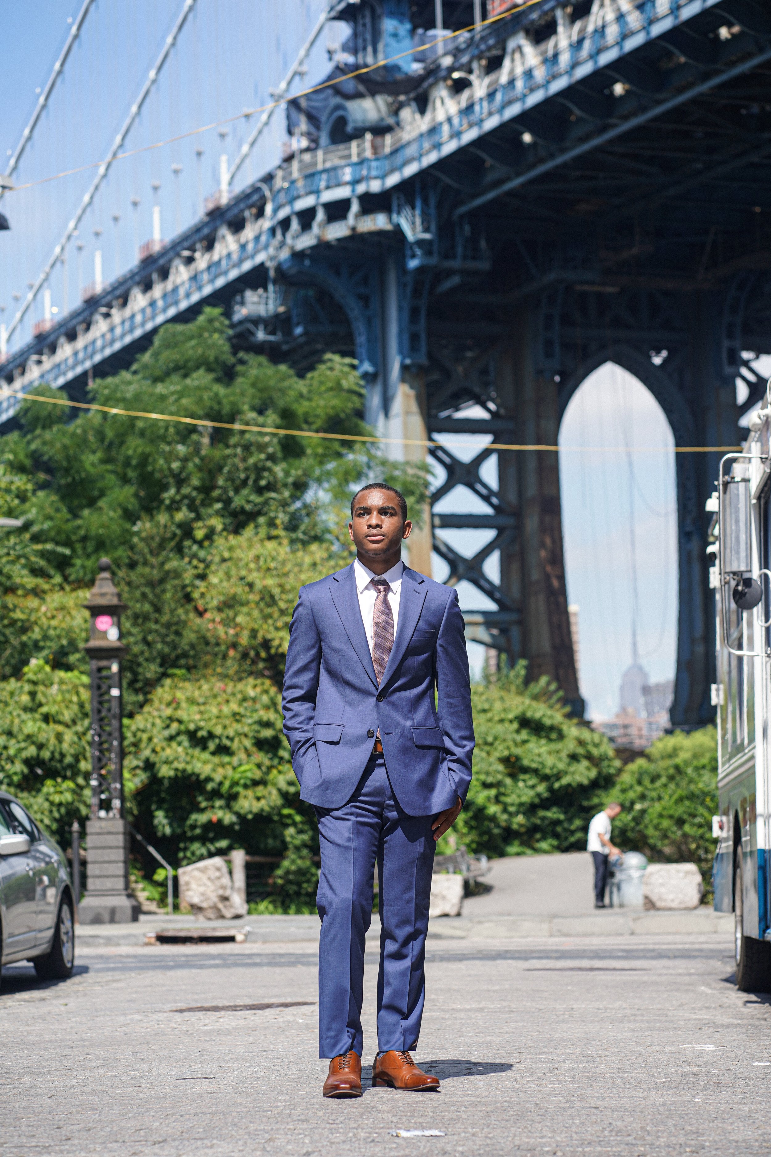 A man in a blue suit walking on a city street with a bridge in the background.