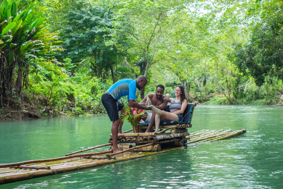 Three people on a bamboo raft in a lush green river with dense trees around, one person is getting on the raft, while the others are seated and smiling.