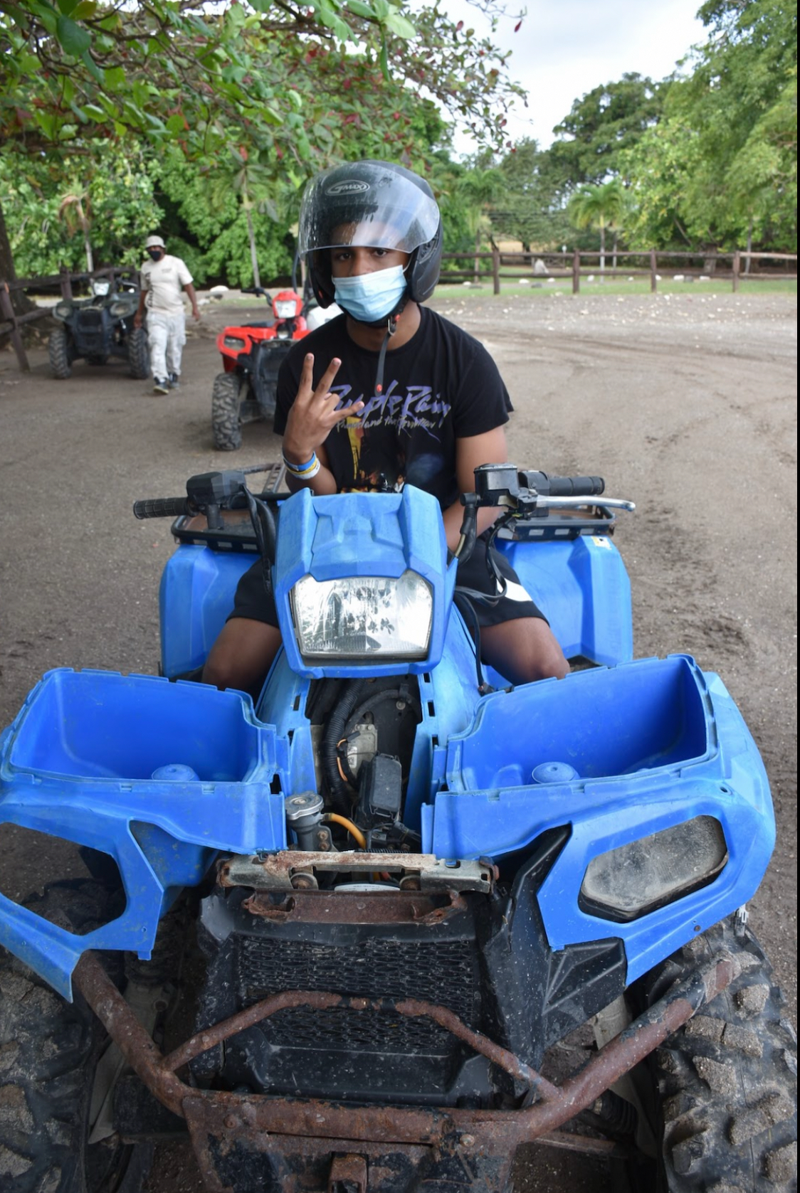 Young person wearing a helmet and face mask sitting on a blue all-terrain vehicle (ATV) outdoors in a natural area with trees.
