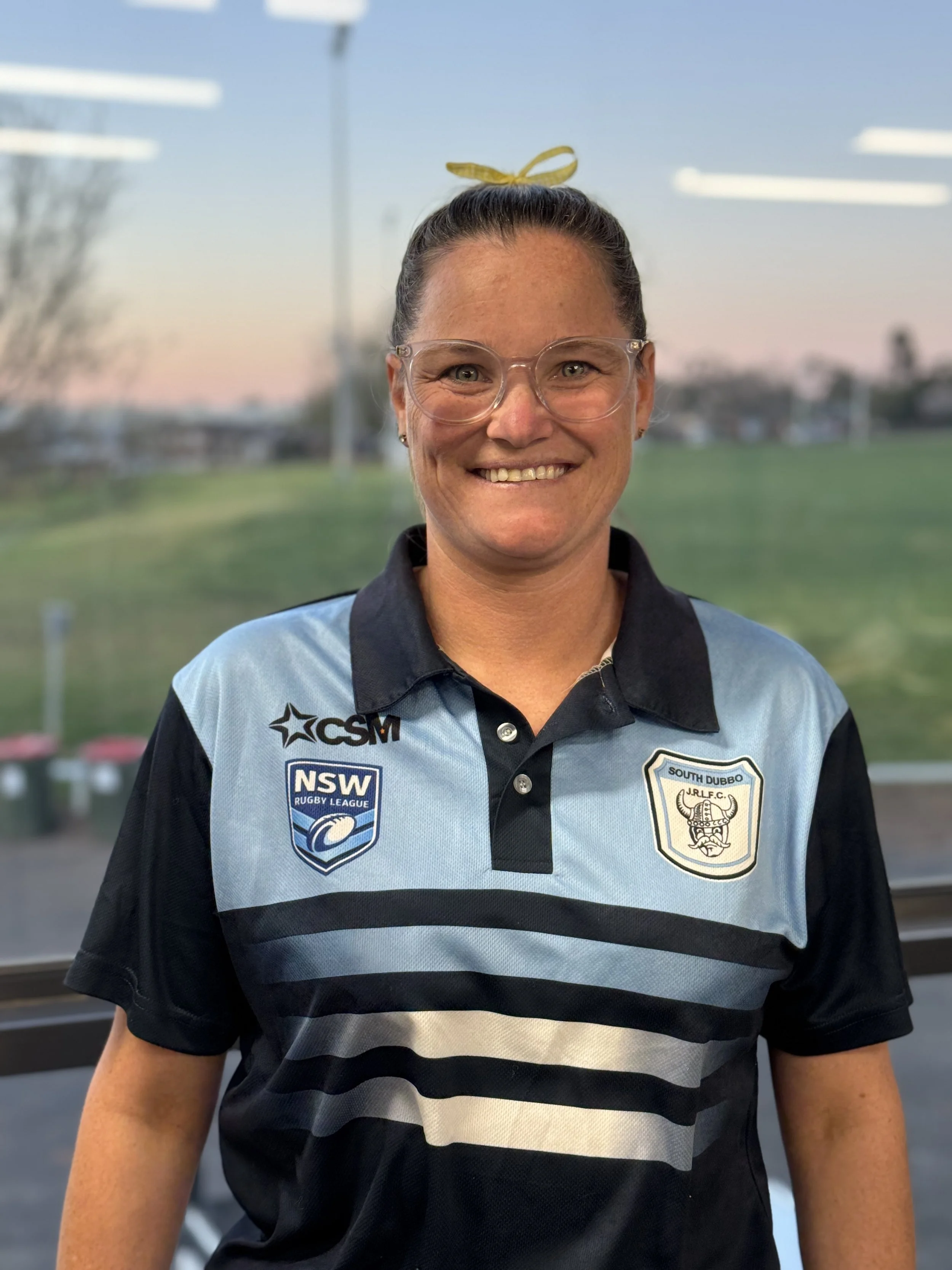 A woman smiling wearing retro-style glasses and a South Dubbo Rugby League jersey, outdoors during sunset or dusk, with a blurred background of a grassy field and trees.
