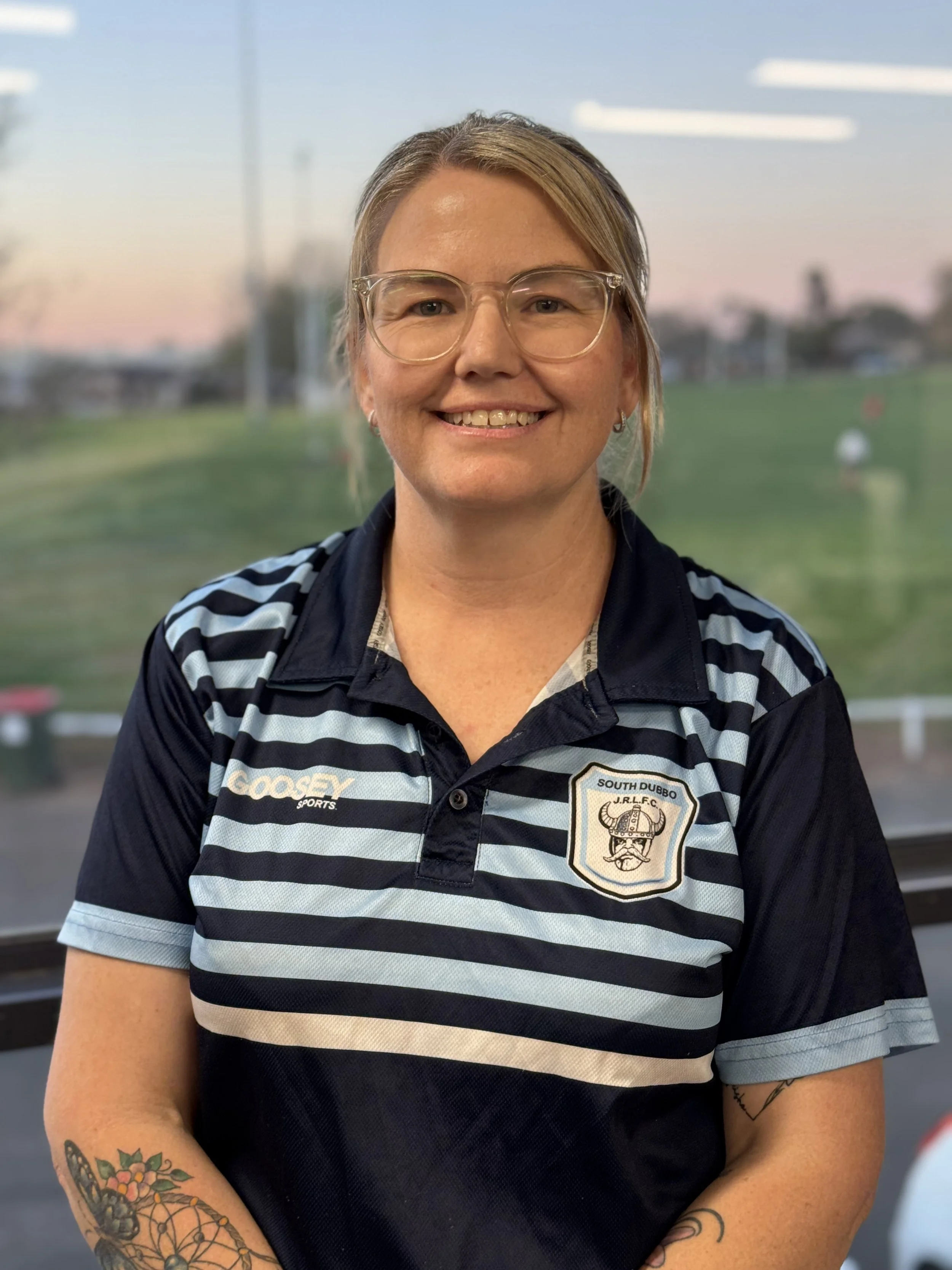 A smiling woman with glasses and tattoos on her arm, wearing a black and white striped golf shirt with a South Dubbo Junior Rugby League Football Club logo, standing outdoors at sunset with a blurred background of a park and people.