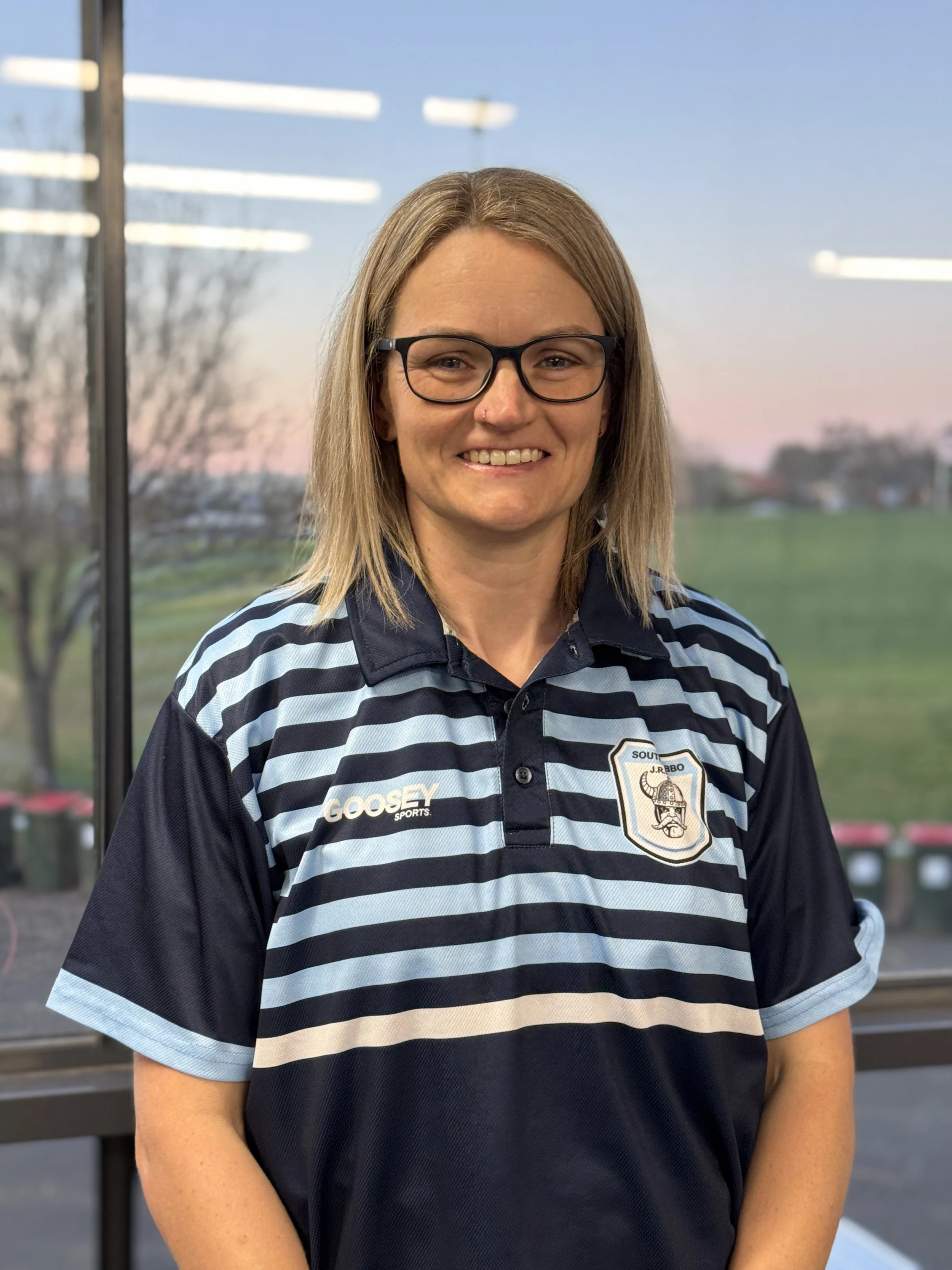 A woman with glasses and shoulder-length blonde hair smiling, wearing a black and light blue striped sports polo with a logo on the chest, standing outdoors with a field, trees, and a sunset sky in the background.
