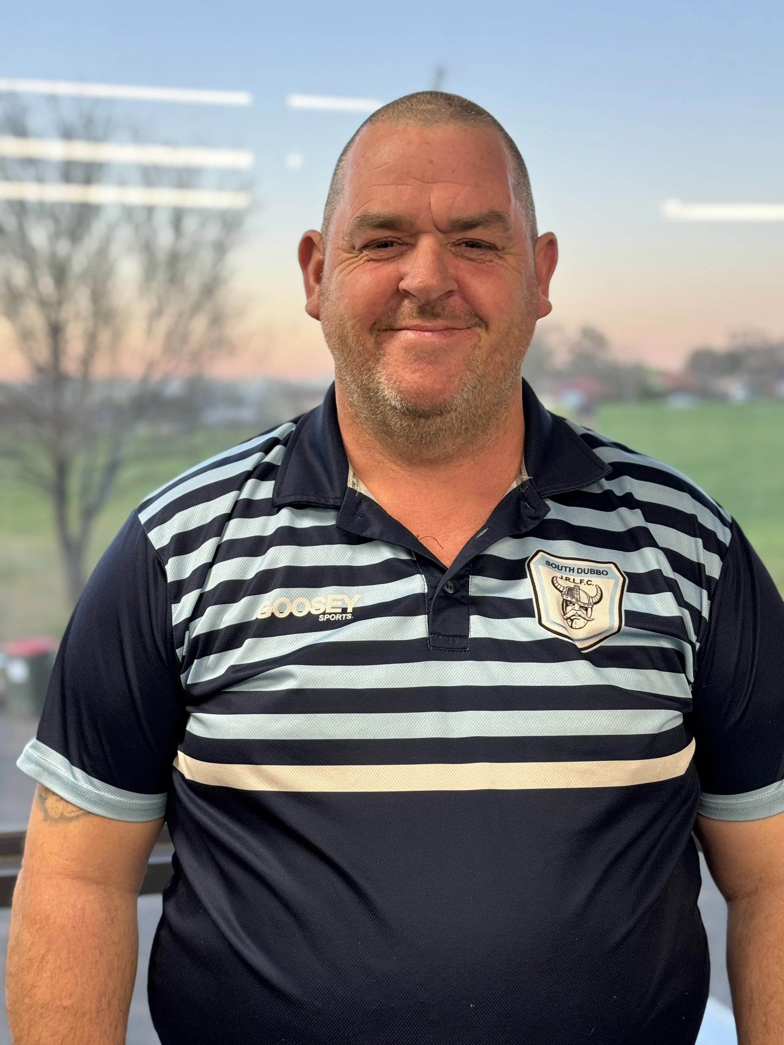A man wearing a blue and white striped sports shirt with an emblem that reads 'South Dubbo Jr. R.L.F.C.' poses outdoors with a blurred background of trees and sky.