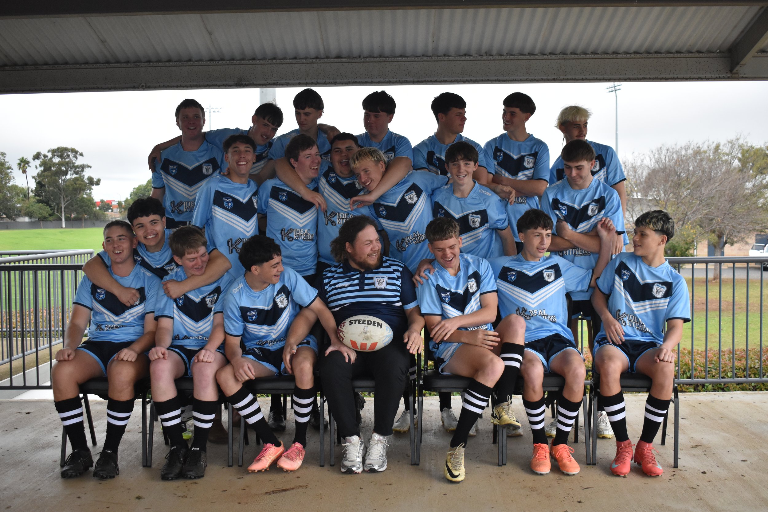 A group of young men in blue sports uniforms sitting and standing on a bench with a rugby ball, posing for a team photo outdoors.