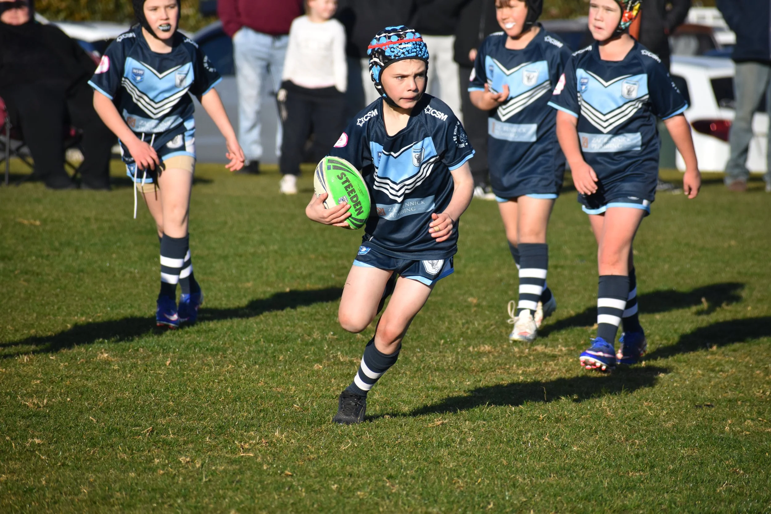A young rugby player running on the field with a green and white rugby ball, wearing a navy blue team uniform with light blue accents and a safety helmet, surrounded by other young players in matching uniforms.