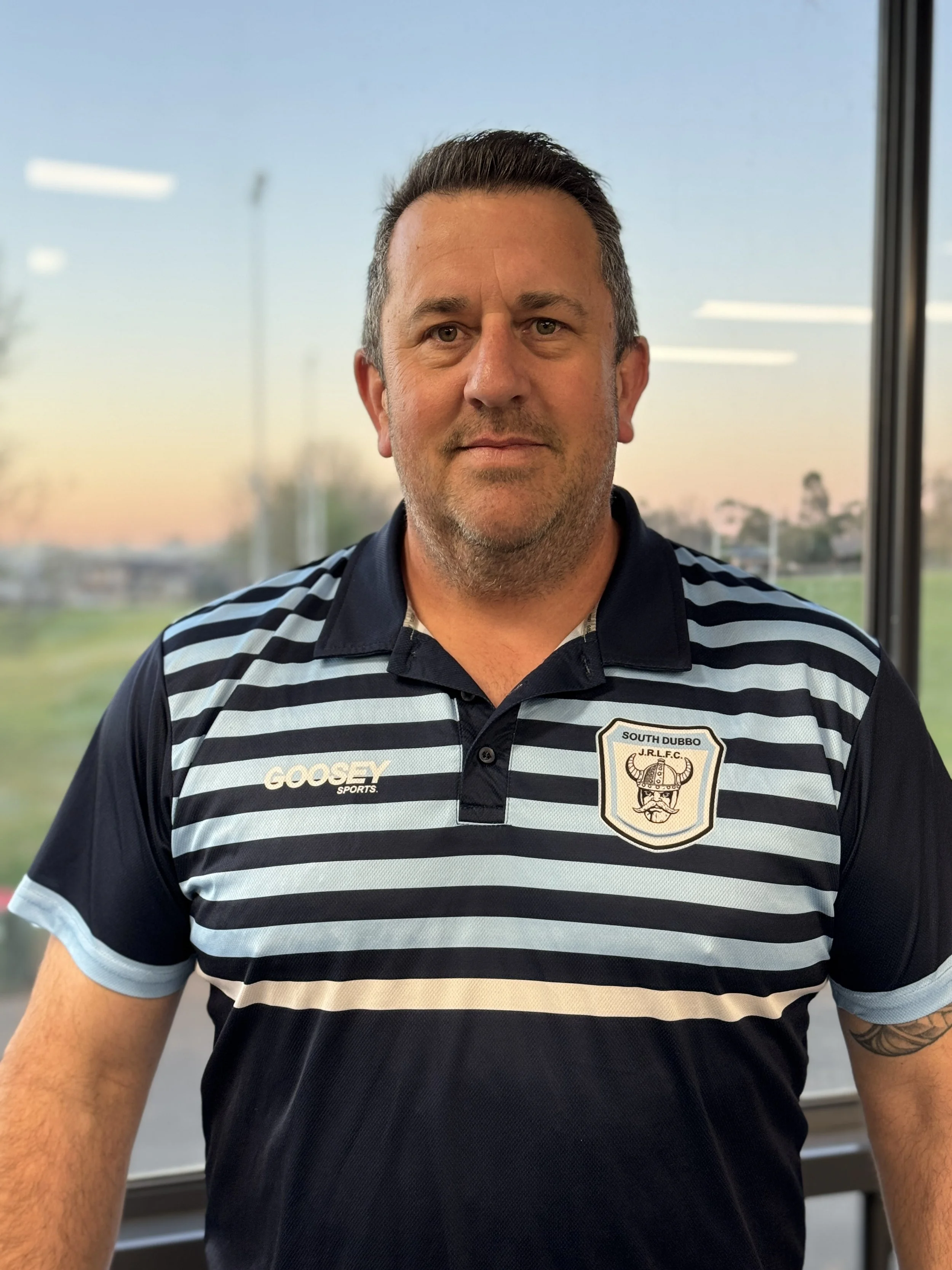A man wearing a navy and white striped polo shirt with a badge that says 'South Dubbo' and 'J.R.L.F.C.' stands indoors near a window, with a blurred outdoor scene in the background.