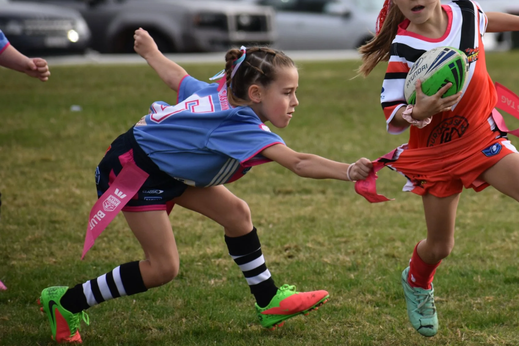 Two young girls are playing rugby outdoors on a grass field. One girl, in a blue and pink uniform, is pulling on the jersey of another girl, who is wearing an orange and black uniform and holding a rugby ball.