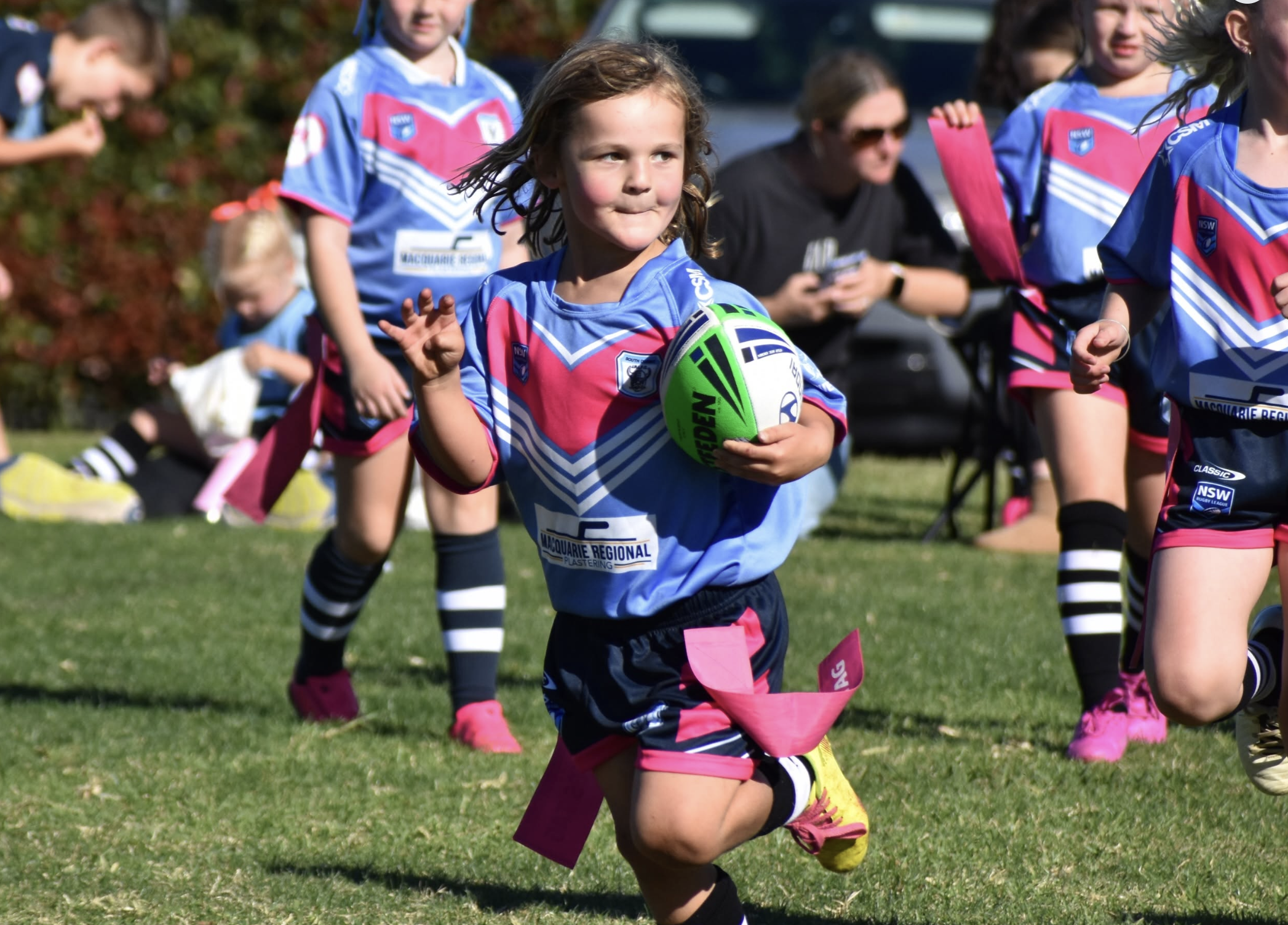Young girl holding a green and white rugby ball running on a grassy field with other children in soccer uniforms.