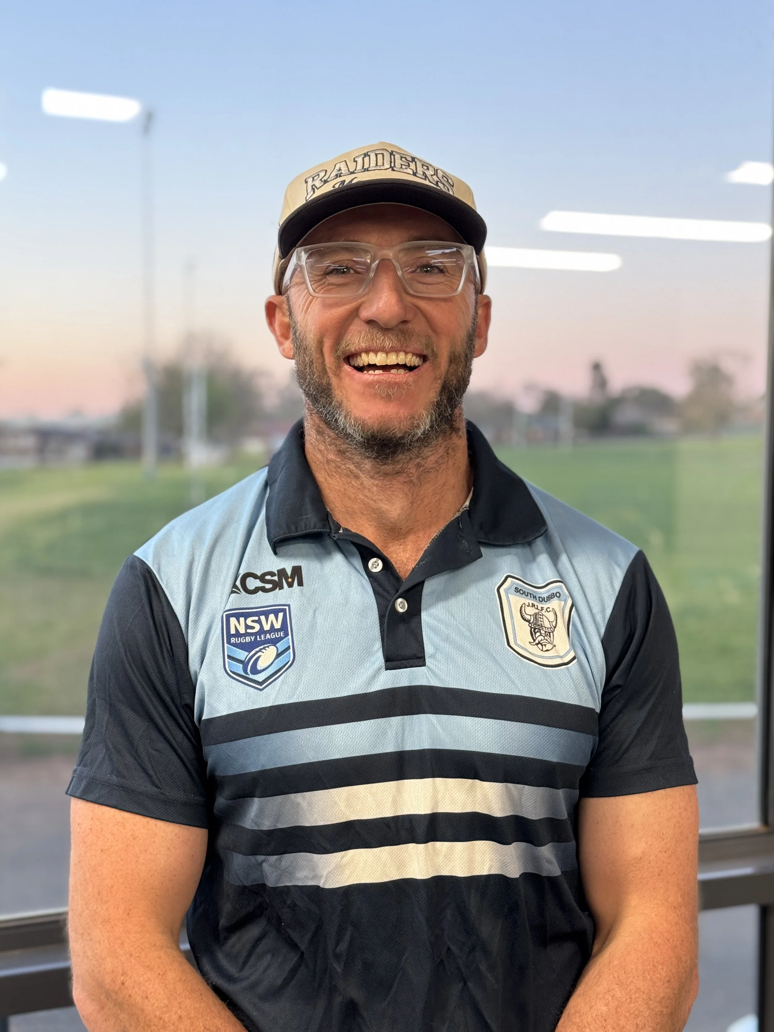 A man with glasses smiling, wearing a beige and black cap with 'RAIDERS' written on it, and a sports shirt with logos, standing outdoors near a sports field.