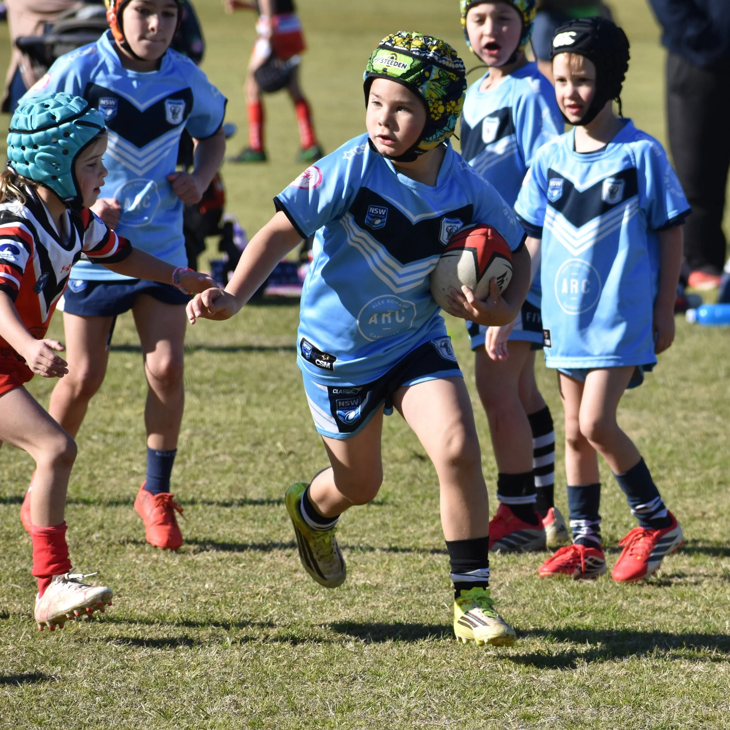 Children wearing protective helmets and jerseys playing rugby on a grassy field, with one child holding a rugby ball.