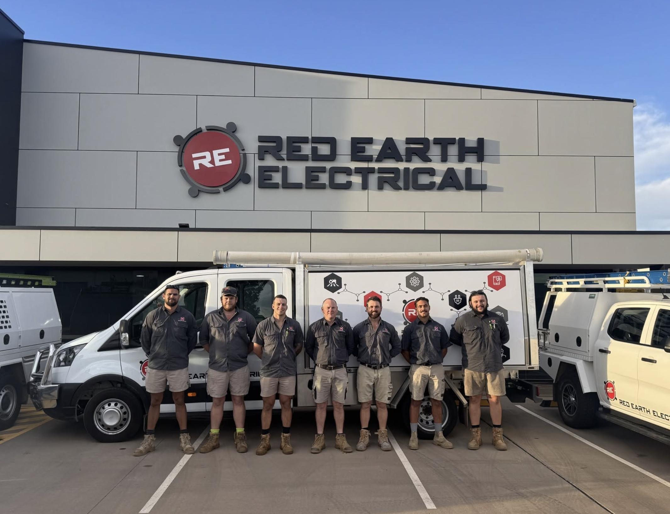 Group of seven workers in uniform standing in front of vehicles and a building with a sign that reads 'Red Earth Electrical'.