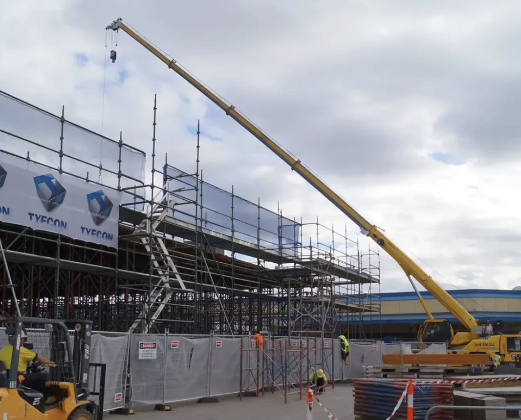 Construction site with scaffolding, a large yellow crane, construction workers wearing safety vests and helmets, and a building under construction with signage that reads 'TYEON' and 'DAISO' in the background.