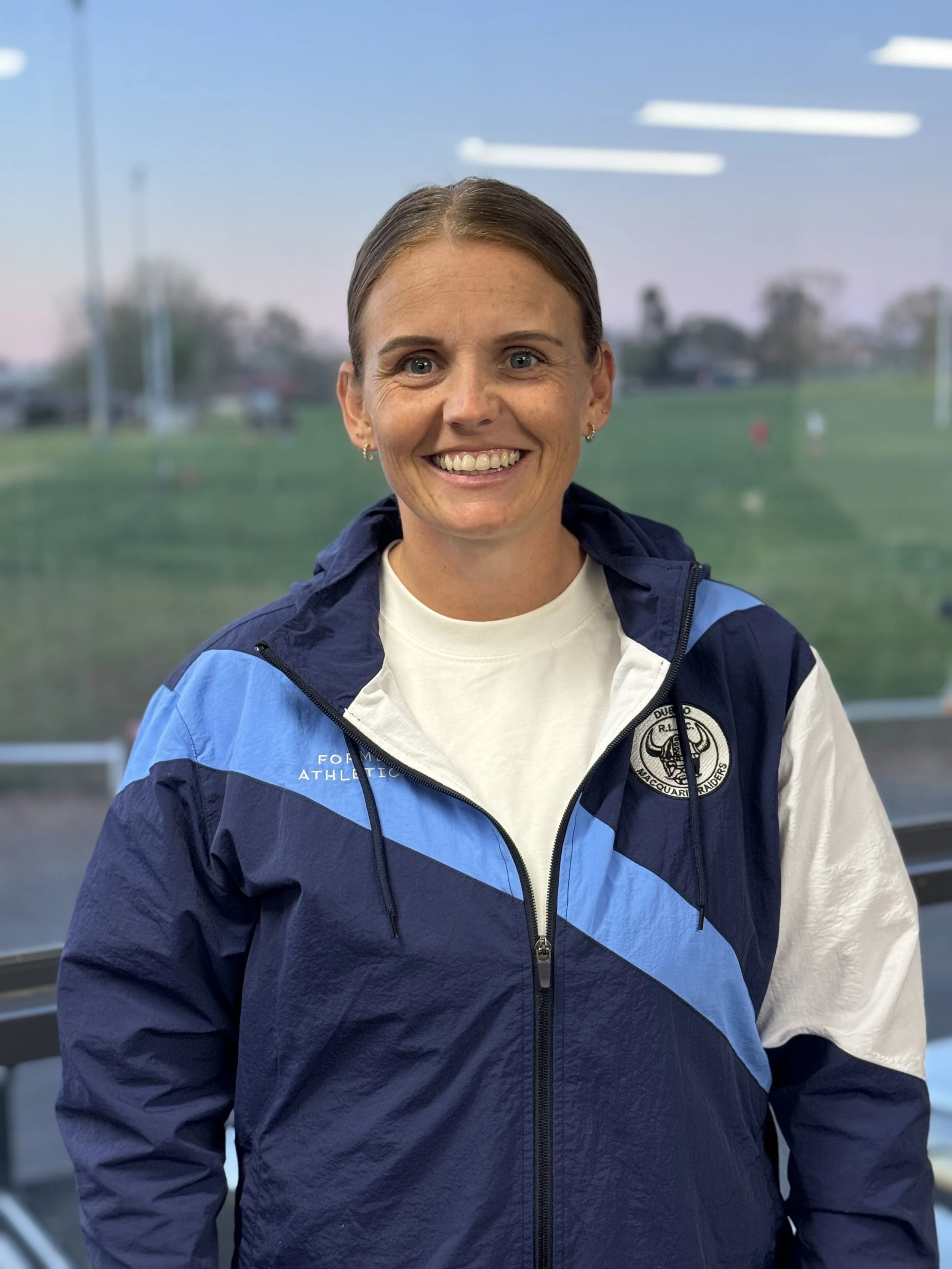 A woman with short brown hair and blue eyes, wearing a white shirt and a blue athletic jacket with a logo, smiling indoors with a large window showing an outside field or park with trees and a blue sky.