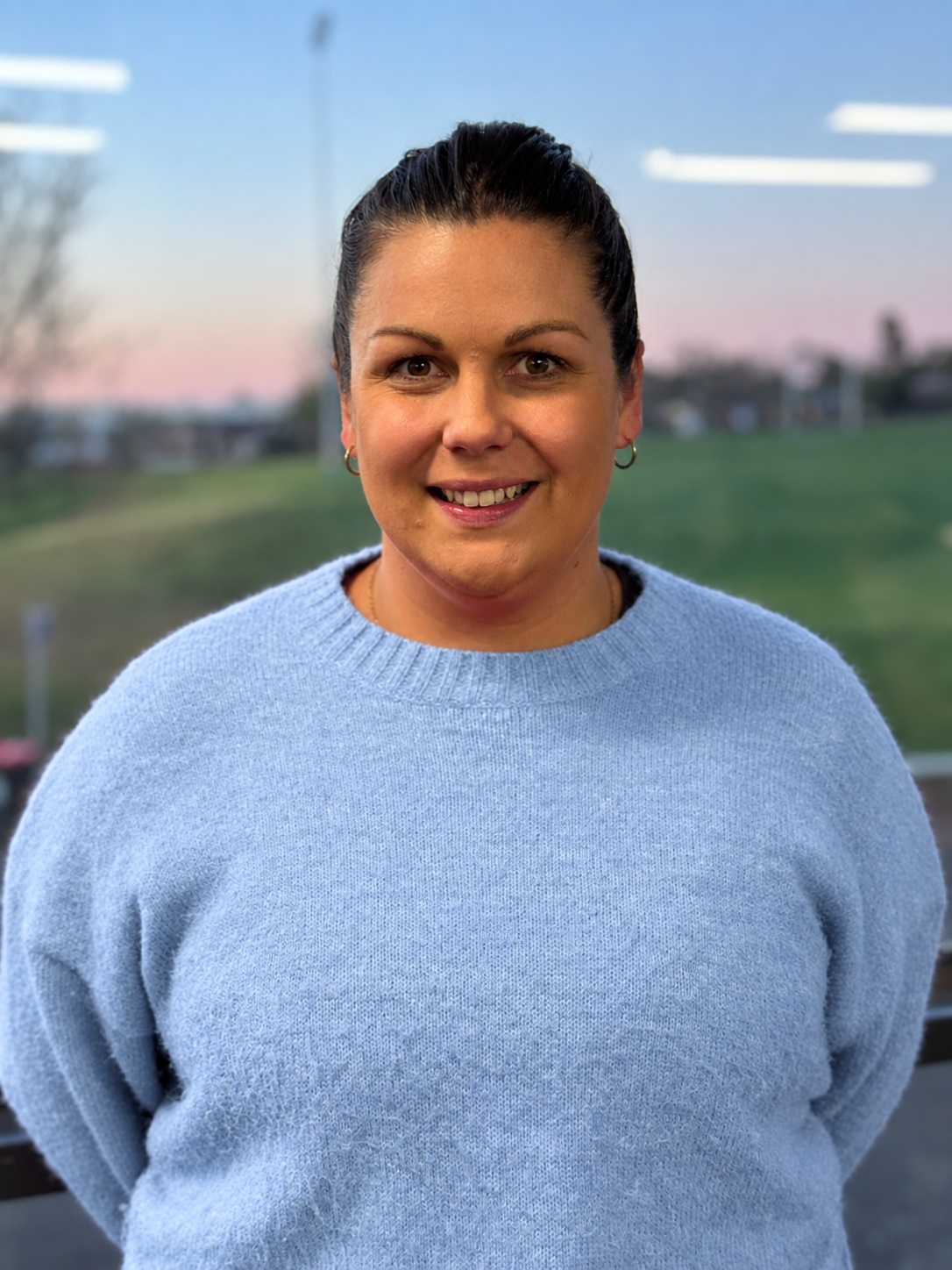 A woman with dark hair pulled back, smiling, wearing hoop earrings and a light blue sweater, standing outdoors during sunset with a blurred background of trees and grass.