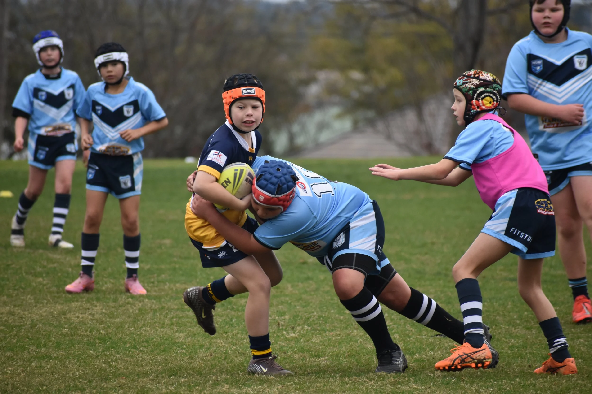 Children playing rugby on a grassy field, with some children tackling and others observing, dressed in sports uniforms with helmets.