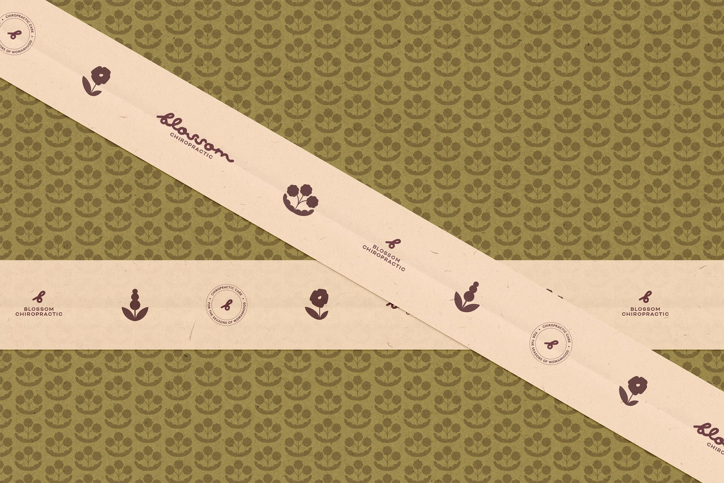 Close-up of a decorative paper with a repeating pattern of flowers and leaves in olive green color. A beige strip running diagonally across the paper features the logo and name of Blossom Chiropractic, along with a small flower icon, in dark purple.