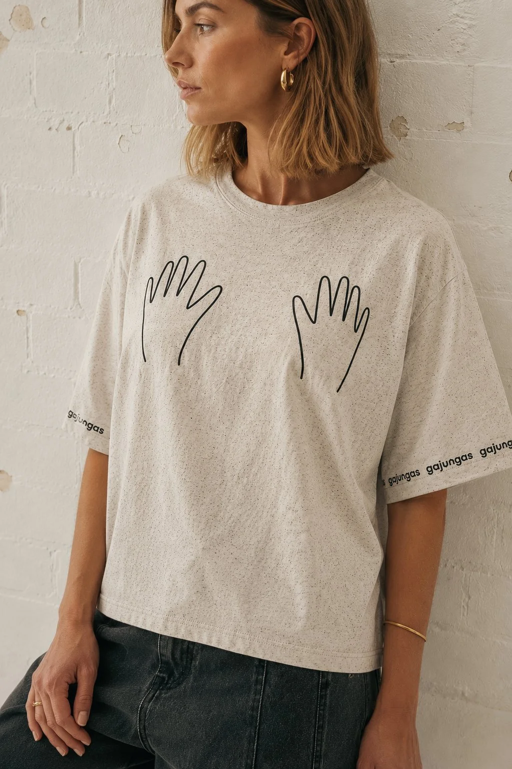A woman with shoulder-length hair wearing a white t-shirt with a black outline drawing of two hands and the text 'gajungas' on the sleeves, standing against a white brick wall.