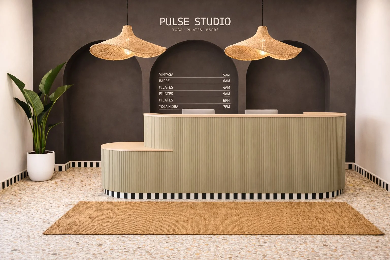 Reception area of Pulse Studio featuring a curved beige reception desk, a potted plant on the left, two hanging woven pendant lights, a dark accent wall with the studio's name and schedule, and a textured beige floor with a rectangular doormat.