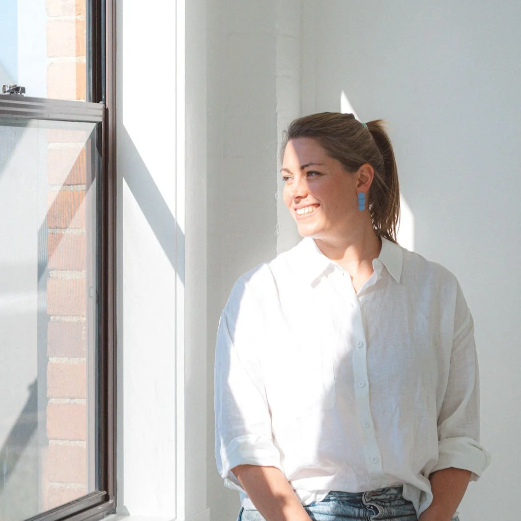 A woman with brown hair tied in a ponytail, wearing blue earrings and a white shirt, standing by a window and smiling while looking outside.