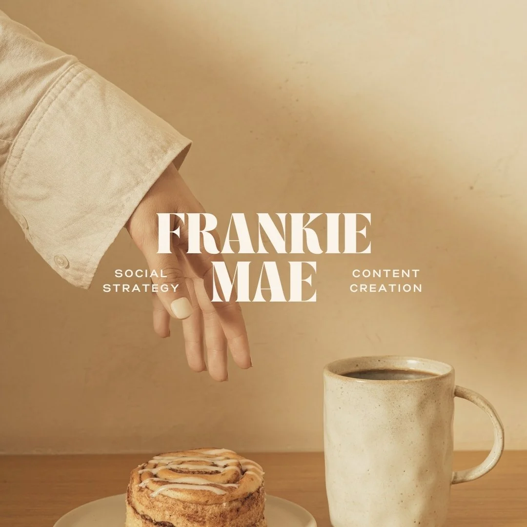 A hand with a beige cuff reaching towards a cinnamon roll with icing on a white plate, next to a mug of coffee on a wooden surface with a beige background.