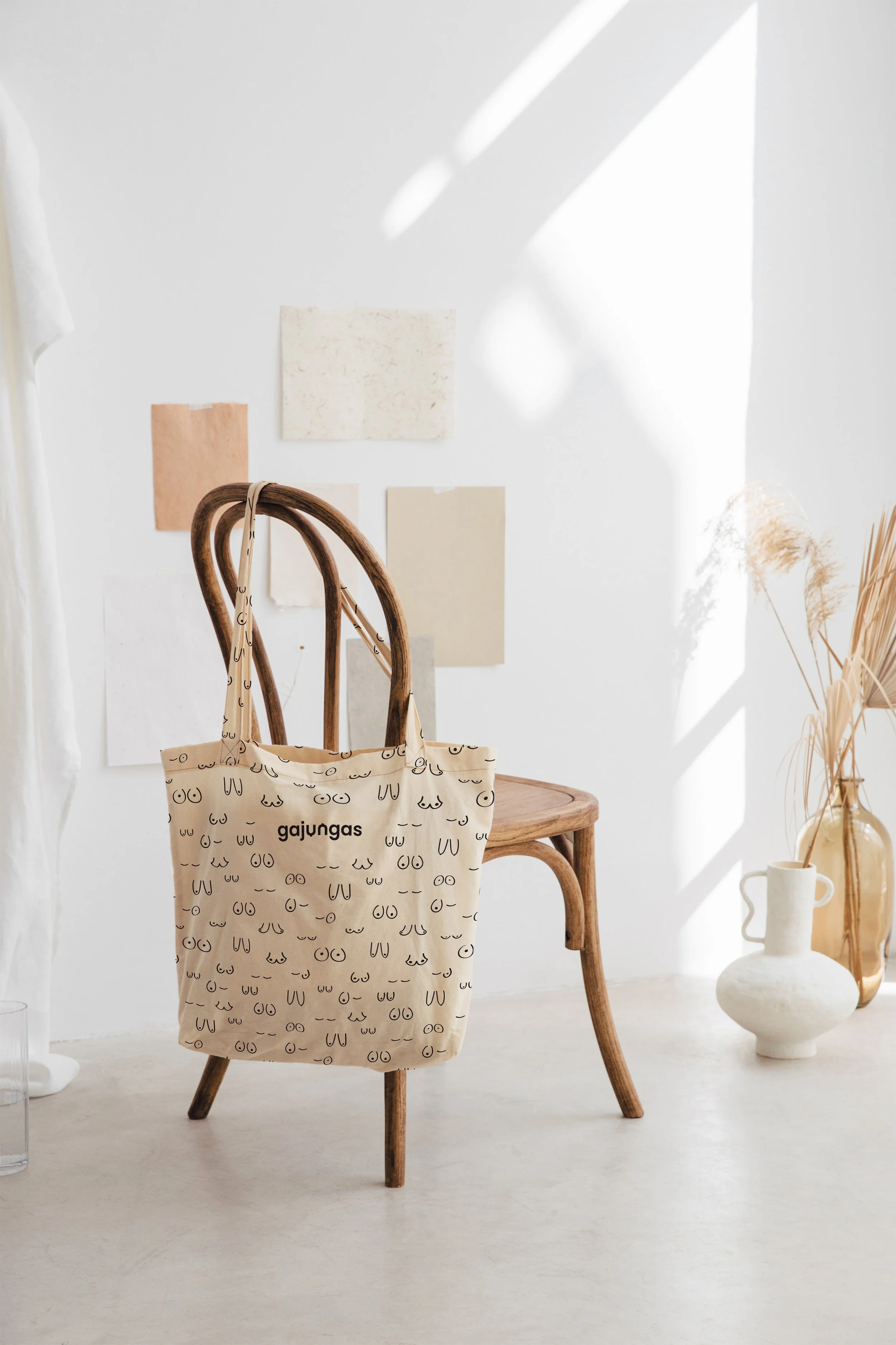 A beige tote bag with black line drawings of faces hangs on the back of a wooden chair in a bright, minimalist room. The room has white walls with abstract color swatches and framed art, and natural light casting shadows. Dried grasses are in a white vase, and a textured ceramic pot is on the floor.