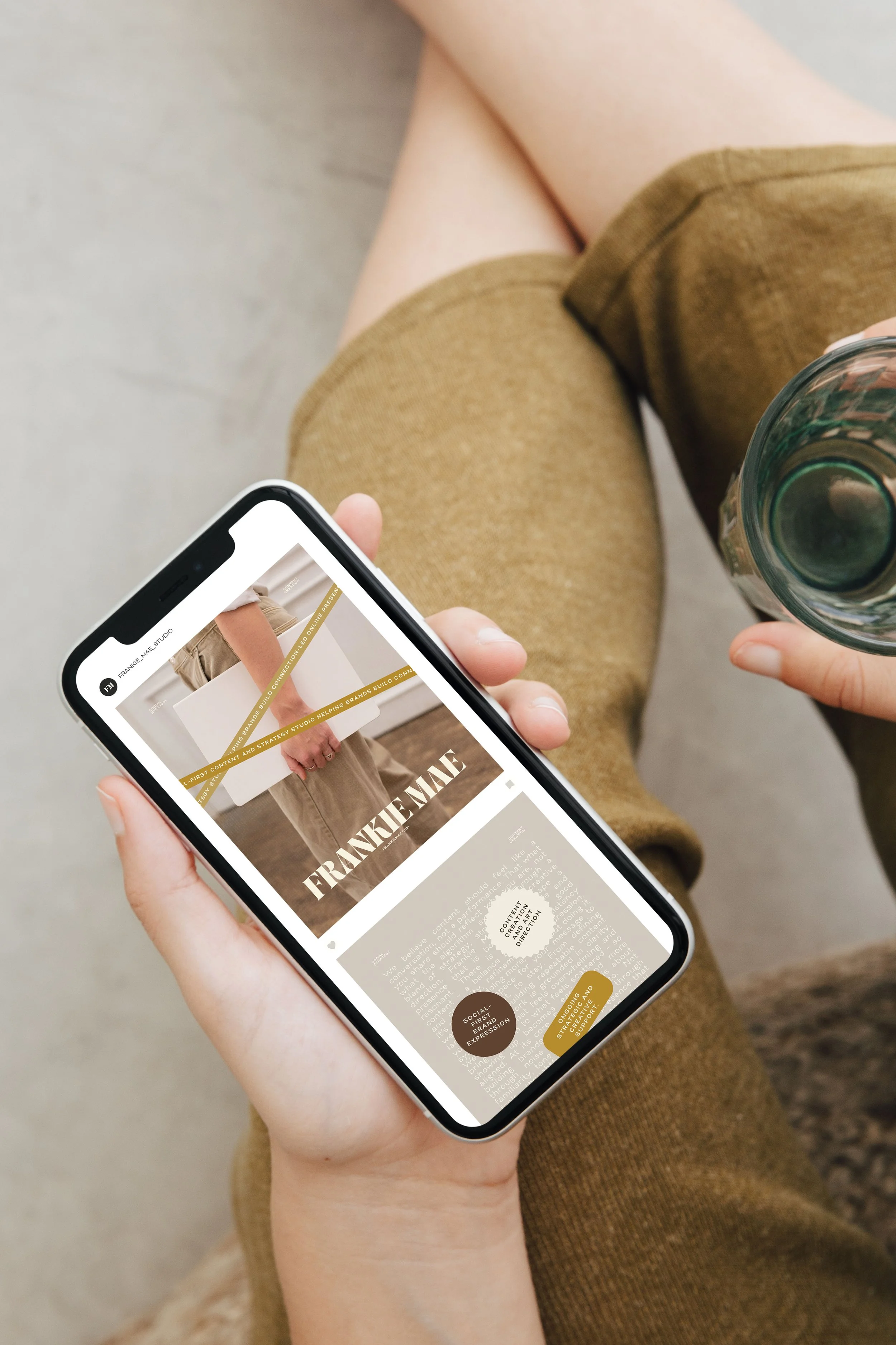 Person holding a smartphone with website content showing a person in a beige outfit and text reading "Frankie Mae." The person is also holding a glass of water, wearing brown shorts, and sitting on a surface with a gray background.