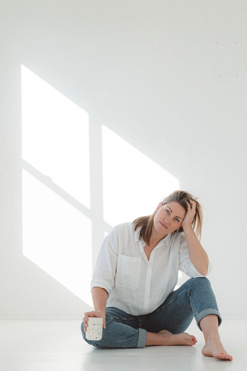 A woman sitting on the floor with one leg bent, holding a cup, in a white room with sunlight streaming through a window casting shadows.