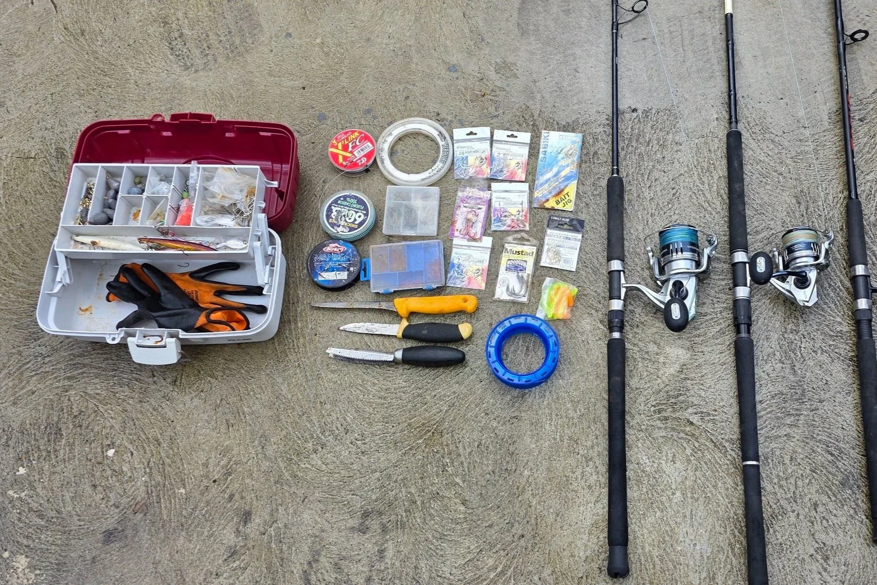 Three fishing rods with reels, fishing tackle boxes containing hooks, weights, and lures, and various fishing accessories and tools such as knives, a screwdriver, and socket rings laid out on a concrete surface.