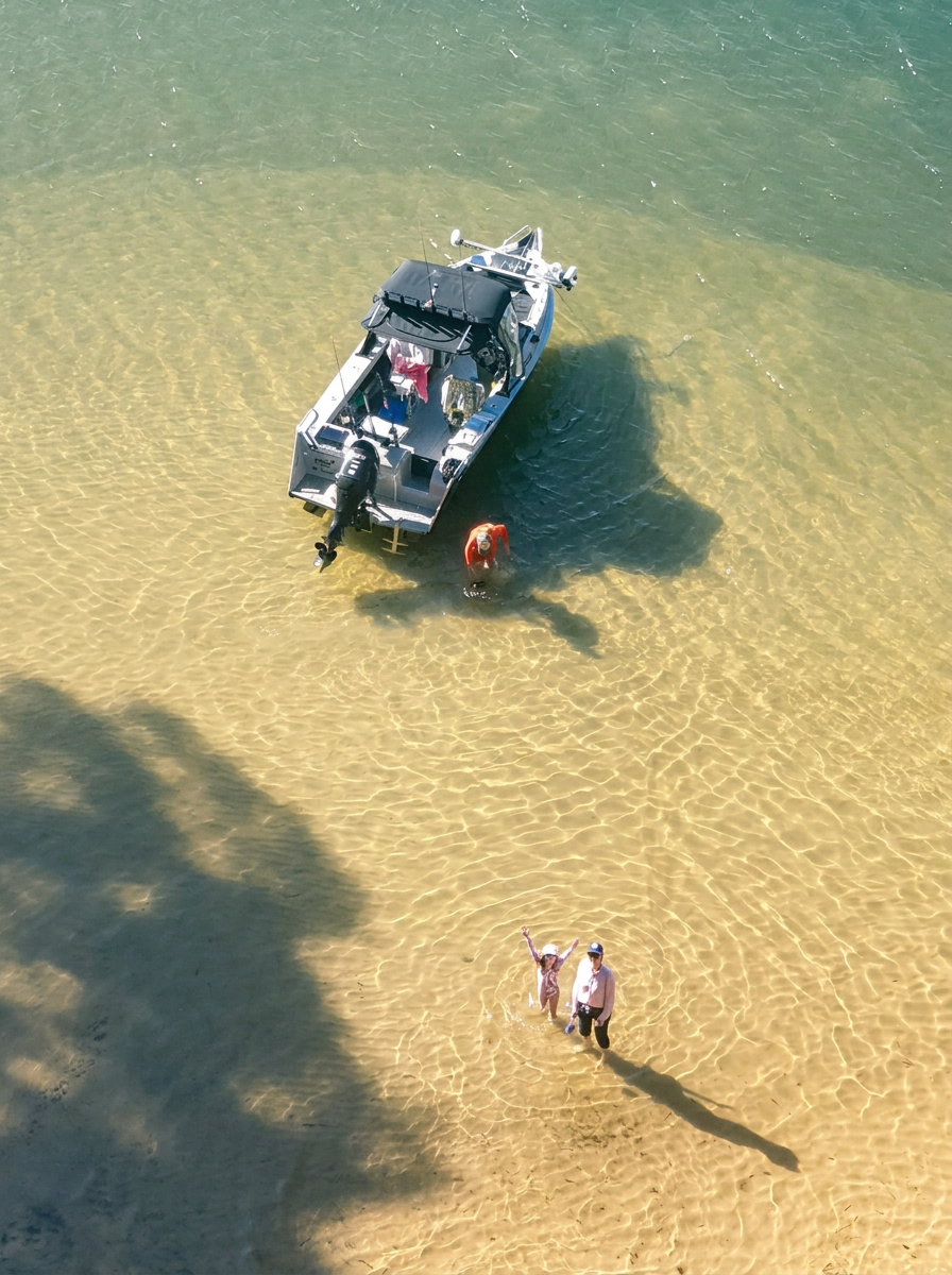 An aerial view of a boat anchored in shallow water near a sandy beach, with a person in an orange shirt standing nearby and two people standing in the water, one of whom is a child waving.