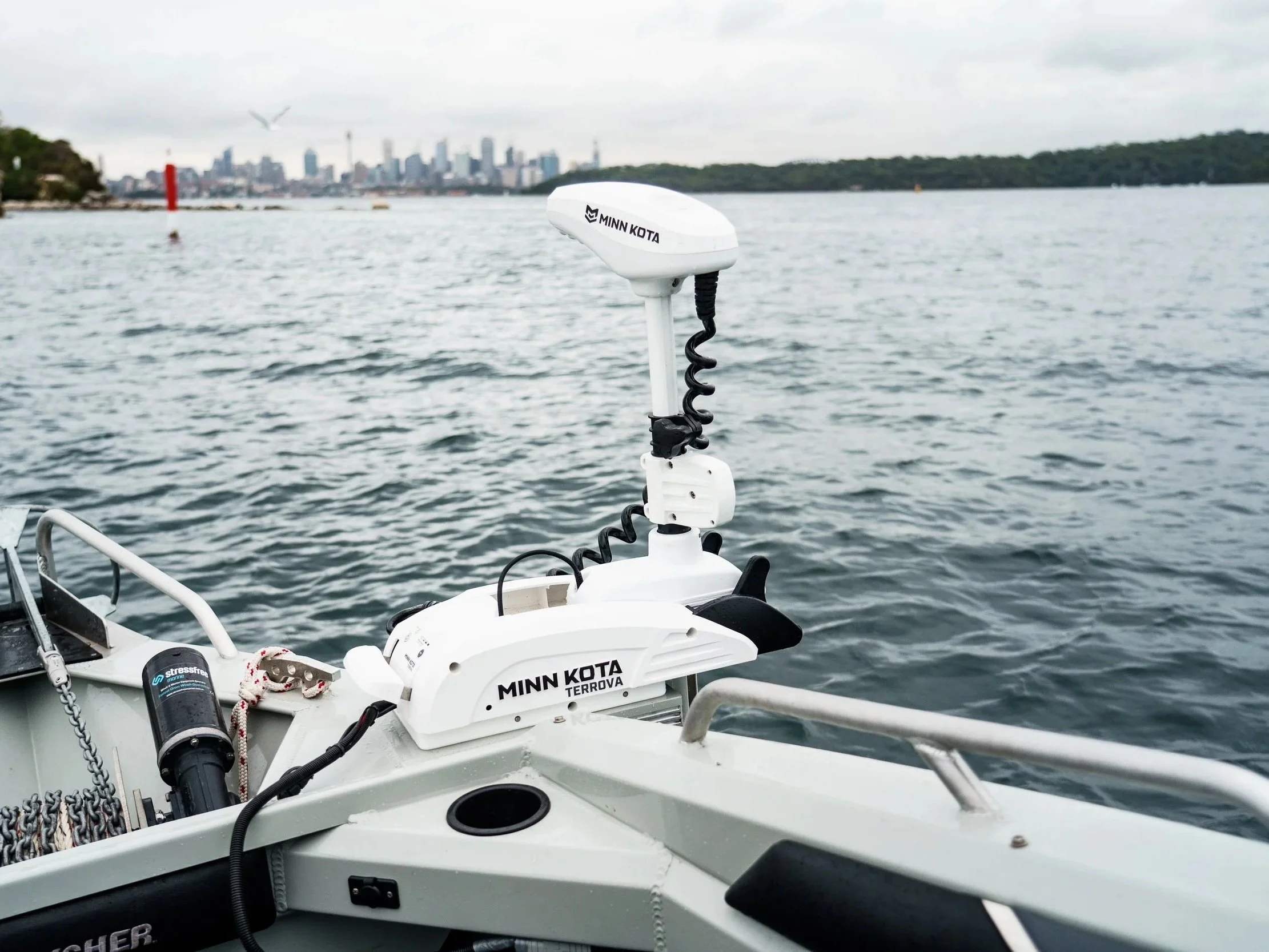 A Minn Kota Terrova trolling motor mounted on a boat with a city skyline and water in the background, under an overcast sky.