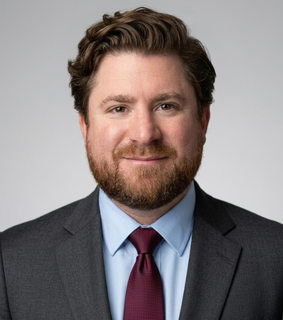 A man with dark wavy hair, a beard, dressed in a gray suit, light blue shirt, and maroon tie, smiling against a plain gray background.