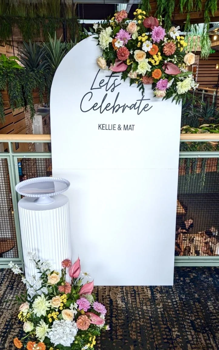 A decorated celebration sign with the message 'Let's Celebrate' for Kellie & Mat, adorned with pink, peach, white, and yellow flowers, situated beside a white pedestal with a floral arrangement, in an indoor venue with lush greenery and wooden accent