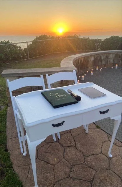 A white writing desk with two chairs, a book titled 'Our Vows', a pen, and a tablet, set outdoors at sunset with a view of the ocean and a candle-lit curved patio.