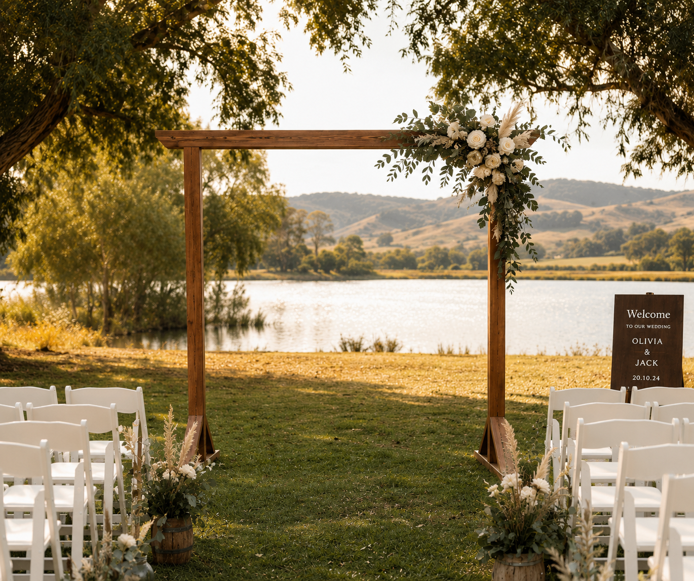 Outdoor wedding setup by a lake with a wooden arch decorated with white flowers and greenery, white chairs on either side, and a welcome sign for Olivia and Jack's wedding, October 24, 2020.