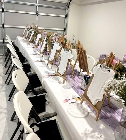 Table set up for a craft or painting activity with easels, framed canvases, and art supplies, decorated with flowers and pink accents.