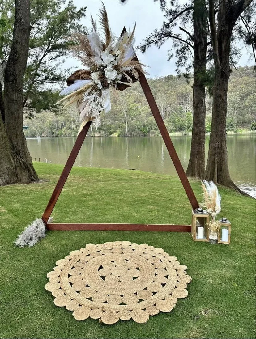 A triangular wooden arch decorated with pampas grass, dried flowers, and white flowers, set on grass near a lake with trees in the background. There are white lanterns and candles on the ground beside it, and a woven circular rug on the grass in fron